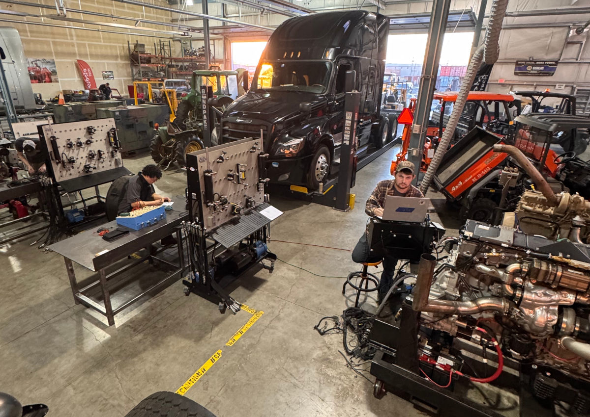 A shot inside the diesel lab. A truck is sitting on the lift. Two students are working at tables, while one student at the right is working on a laptop next to a diesel engine.